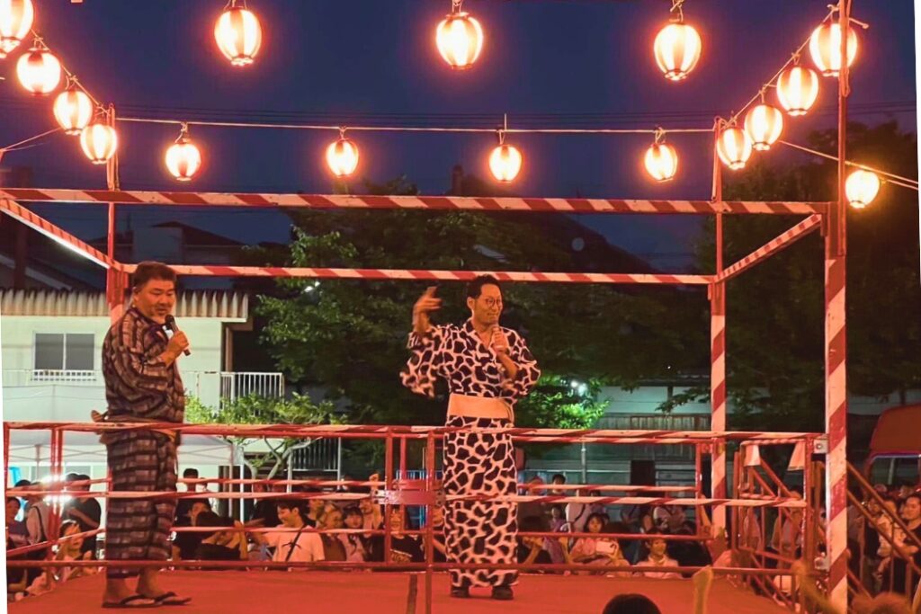 夏祭りの櫓に立ち、浴衣姿でマイクを持つ明山聡一郎さん。地域を盛り上げる活動の一場面。