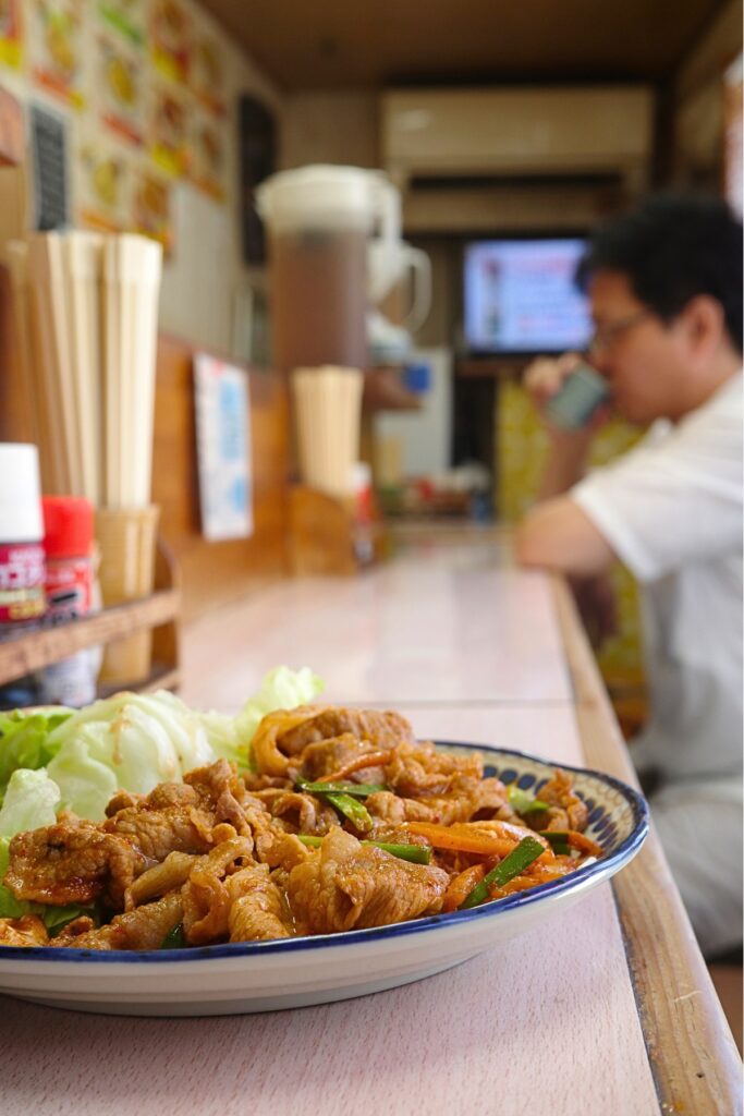 どんぶり亭の豚キムチ定食。カウンター席で食事をする学生と昭和区の定食屋らしい店内風景